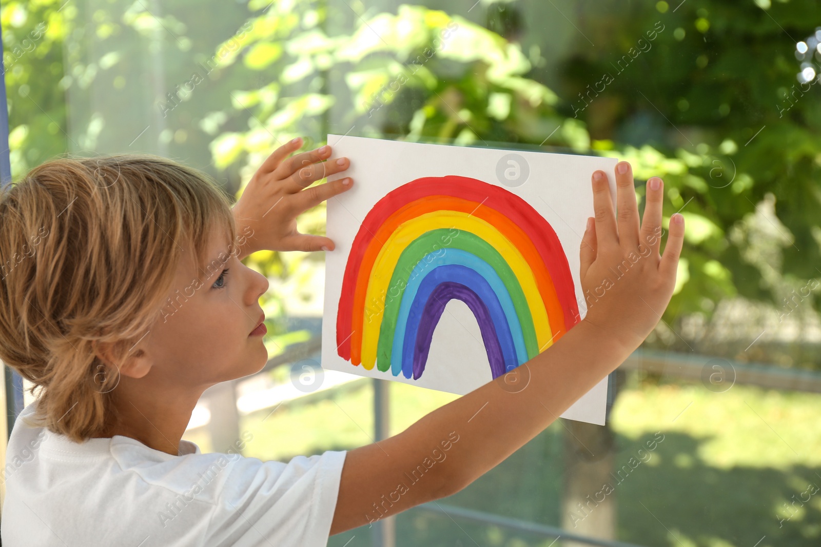 Photo of Little boy holding rainbow painting near window. Stay at home concept