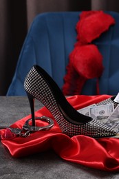 Photo of Prostitution concept. High heeled shoe, dollar banknotes, condom and handcuffs on grey textured table near chair with bra. Space for text