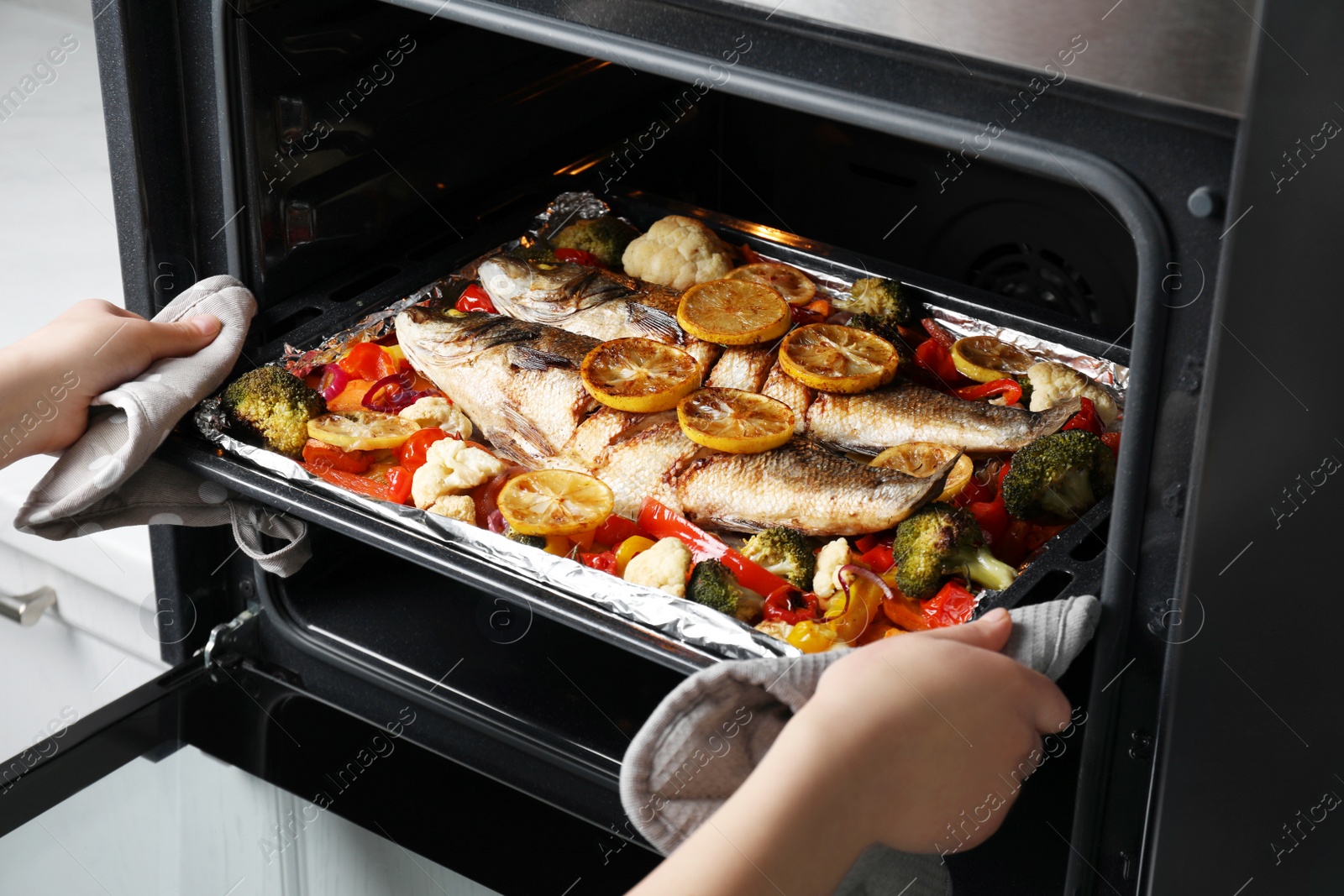 Photo of Woman taking out baking tray with sea bass fish and vegetables from oven, closeup