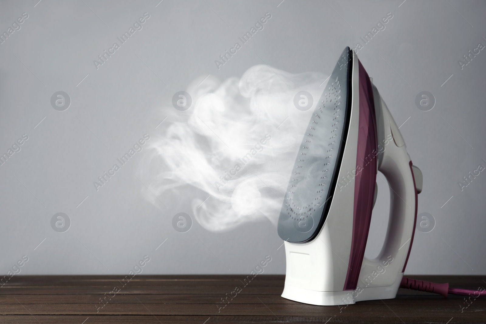 Image of Modern iron with steam on wooden table against light grey background, space for text