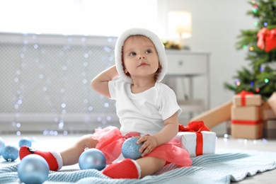 Cute baby in festive costume playing with Christmas decor on floor at home