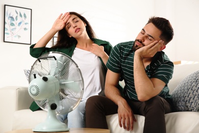 Photo of Couple suffering from heat in front of fan at home. Summer season