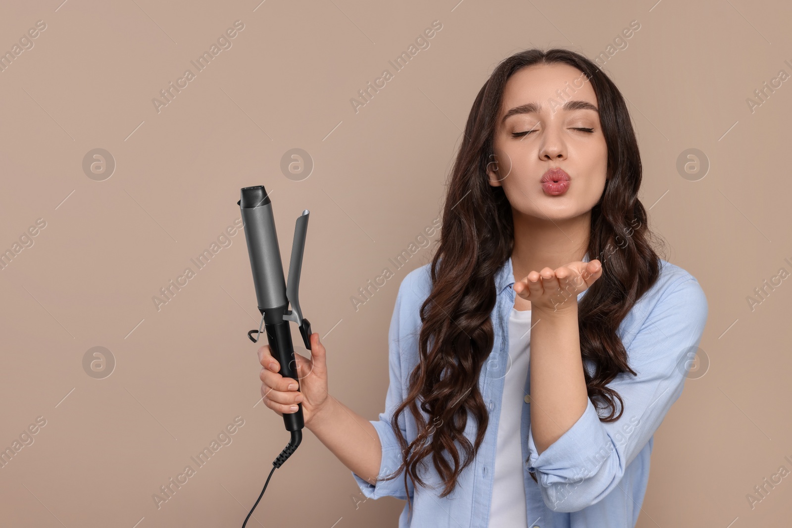 Photo of Beautiful woman with curling hair iron blowing kiss on beige background. Space for text