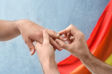 Young man putting wedding ring on his boyfriend's finger near rainbow flag. Gay marriage