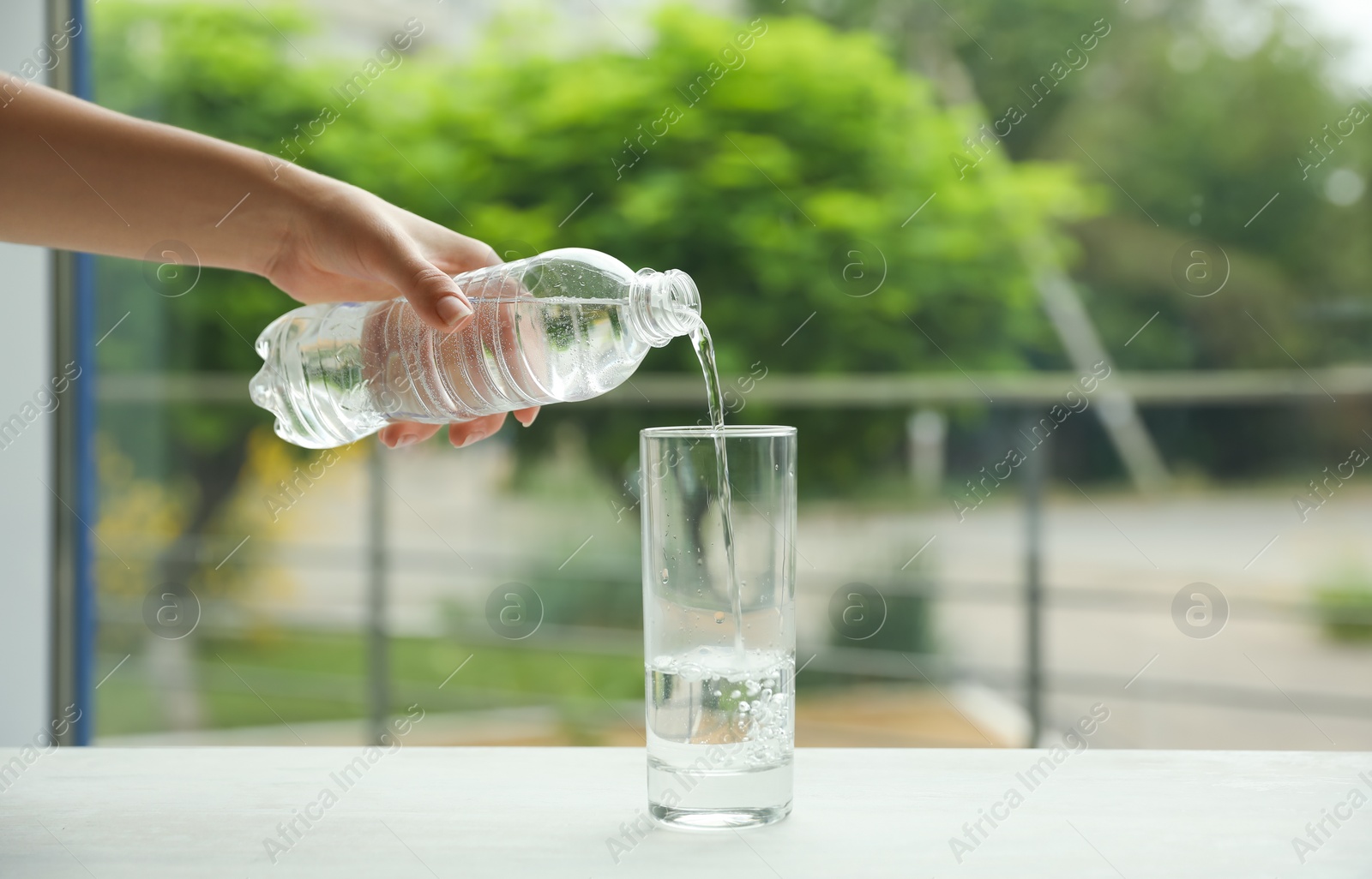 Photo of Woman pouring water from bottle into glass on table against blurred background, closeup