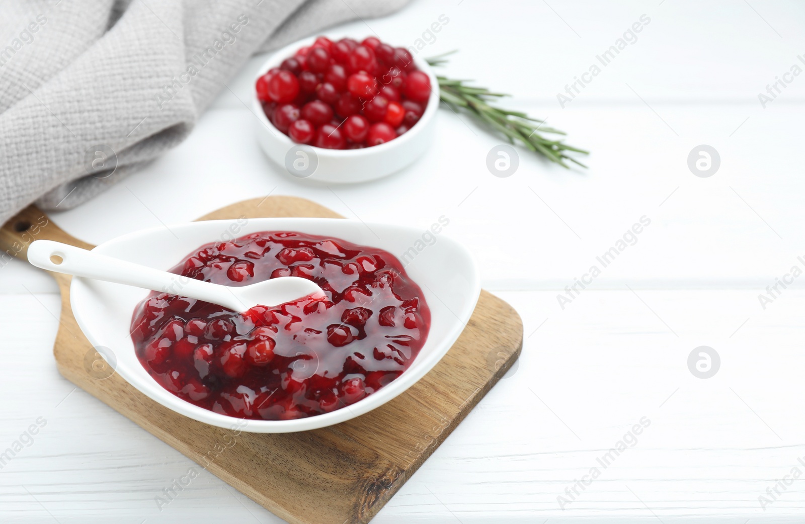 Photo of Tasty cranberry sauce in bowl, fresh berries and rosemary on white wooden table. Space for text