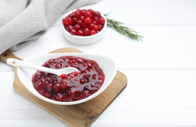 Tasty cranberry sauce in bowl, fresh berries and rosemary on white wooden table. Space for text