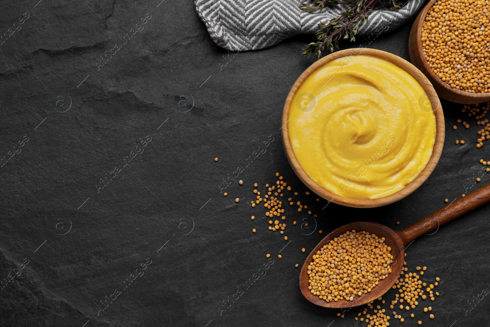 Photo of Bowl with delicious mustard and seeds on black textured table, flat lay. Space for text