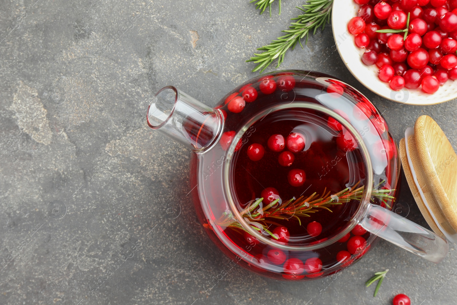 Photo of Tasty hot cranberry tea in teapot, rosemary and fresh berries on light grey textured table