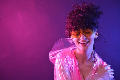 Beautiful young woman in transparent coat and sunglasses posing on color background in neon lights. Space for text