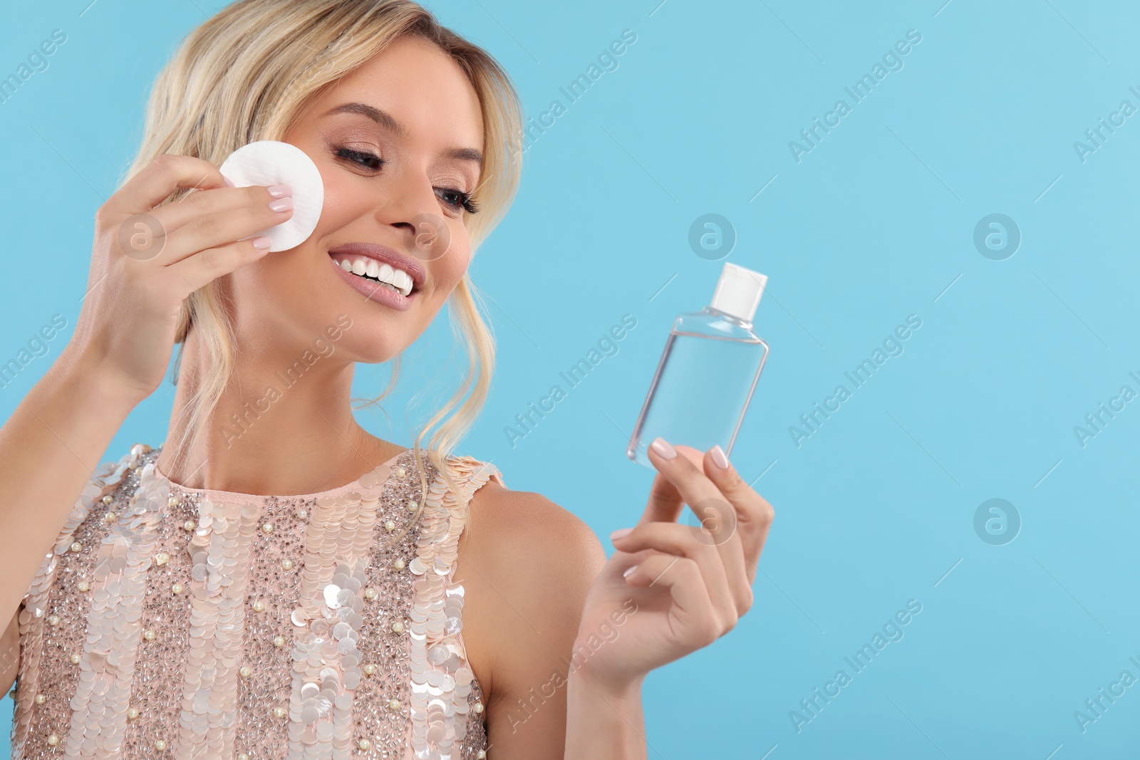 Photo of Smiling woman removing makeup with cotton pad and holding bottle on light blue background. Space for text