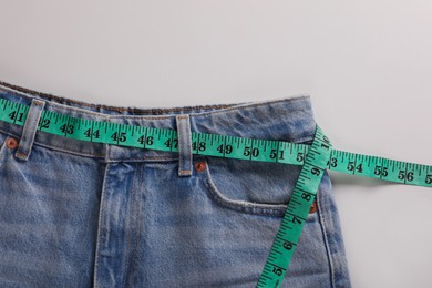 Jeans with measuring tape on light grey background, top view. Weight loss concept