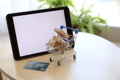 Photo of Internet shopping. Small cart with boxes and credit card near modern tablet on table indoors