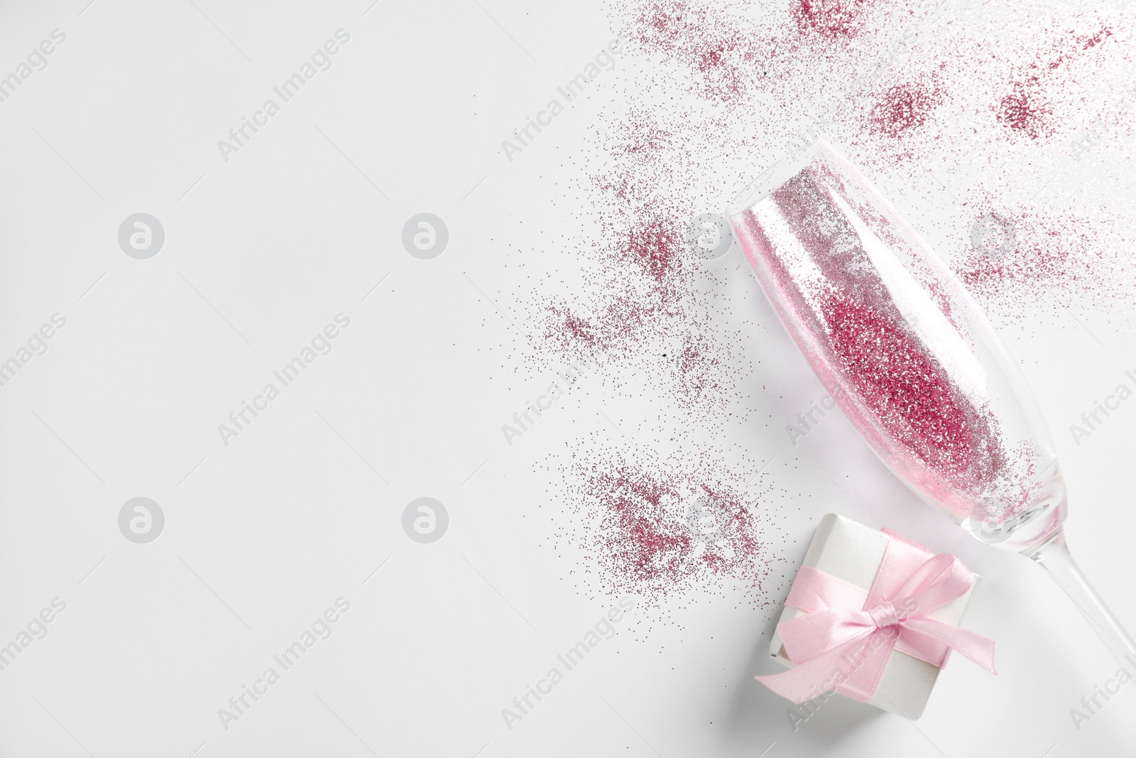 Photo of Champagne glass with pink glitter, gift box and space for text on white background, top view. Hilarious celebration