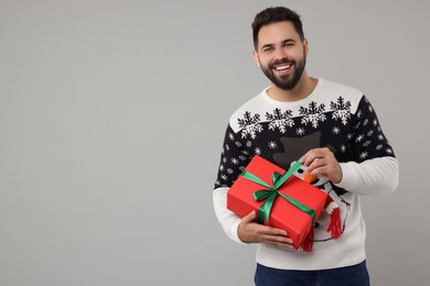 Young man in Christmas sweater opening gift on grey background. Space for text
