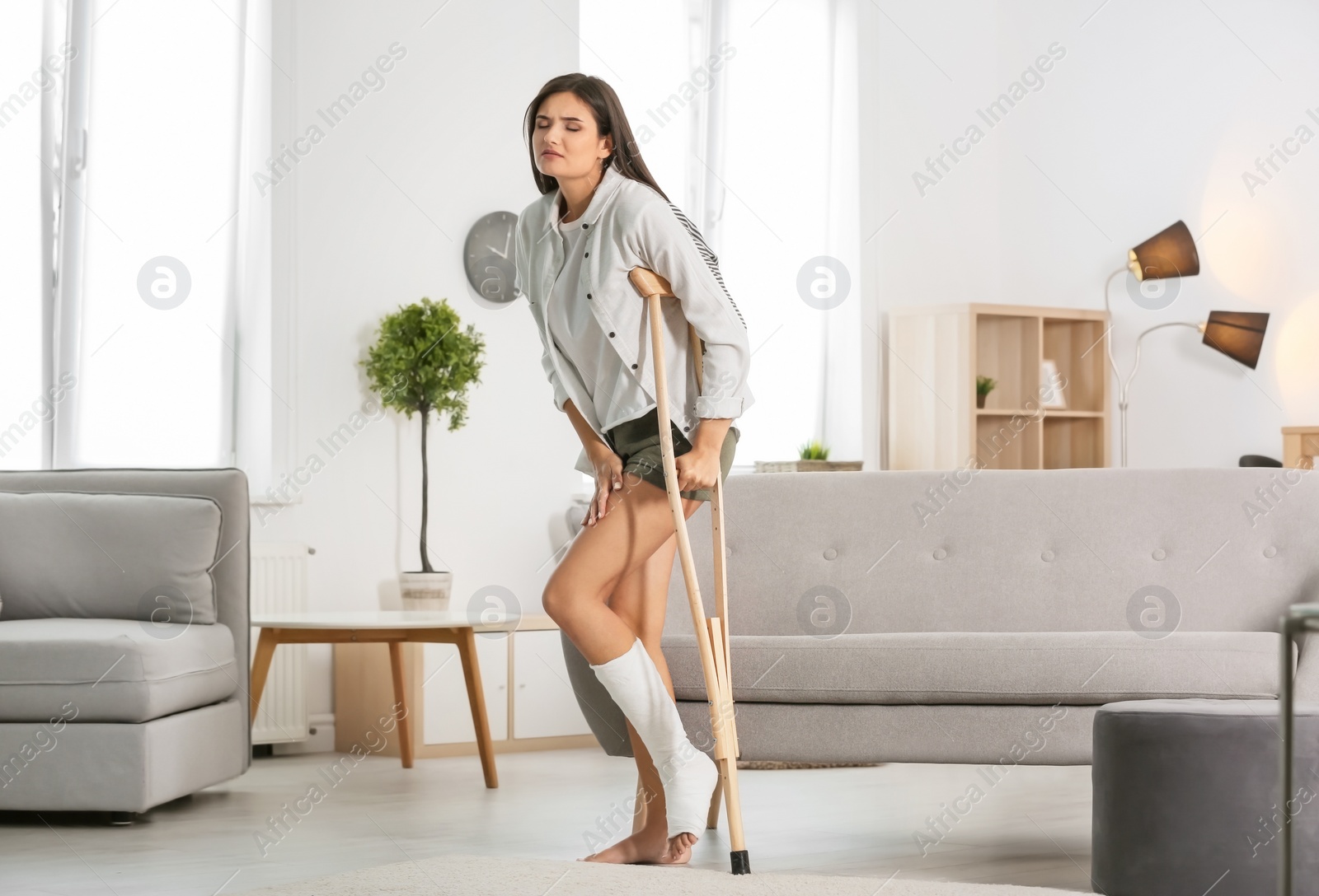 Photo of Young woman with crutch and broken leg in cast at home