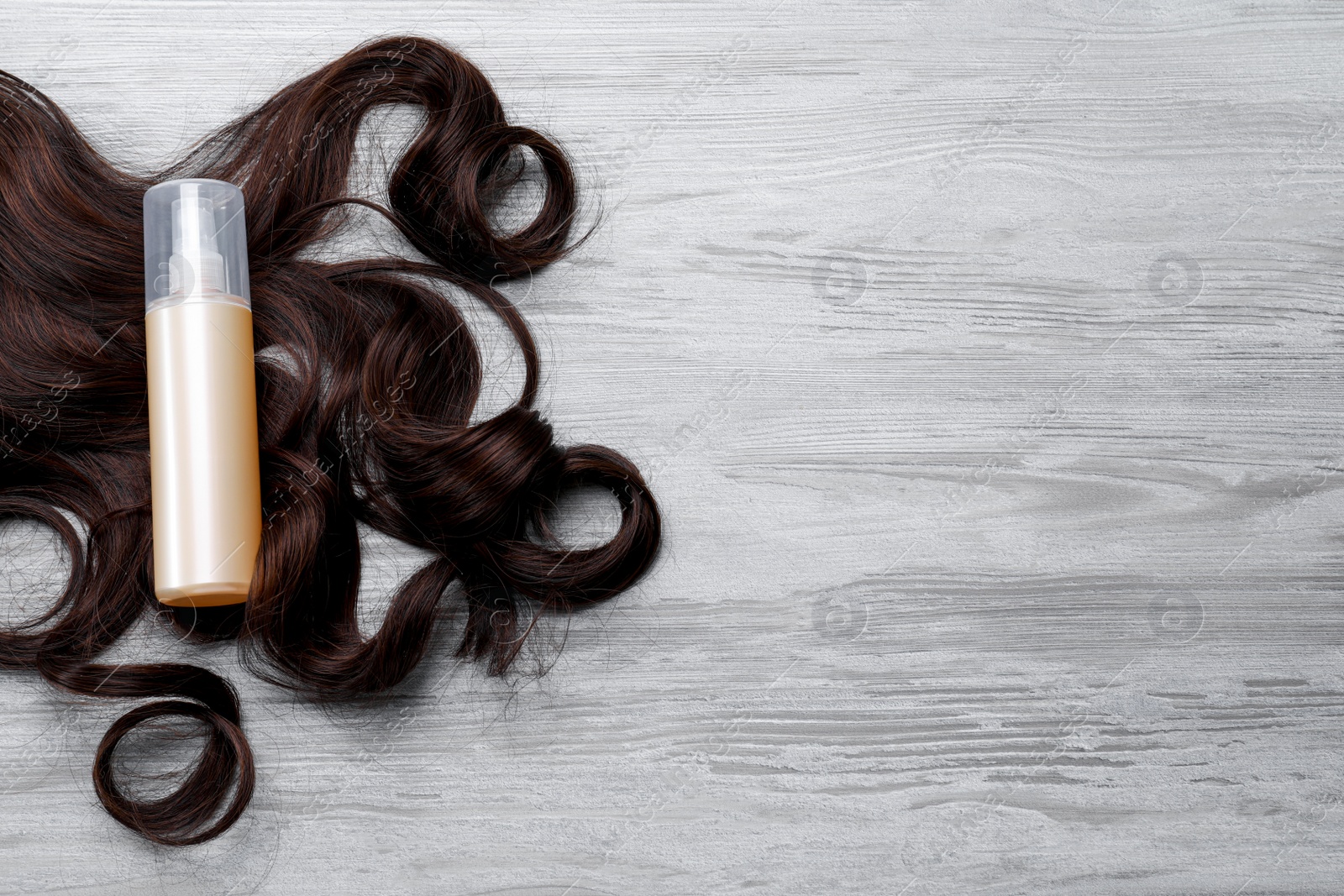 Photo of Spray bottle with thermal protection and lock of brown hair on white wooden table, flat lay. Space for text