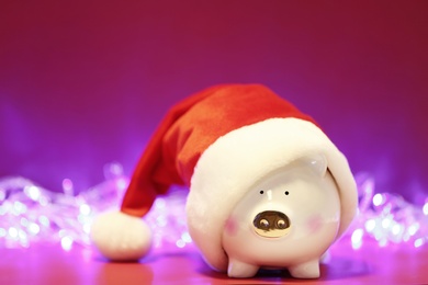 Photo of Piggy bank with Santa hat on table against blurred Christmas lights. Space for text