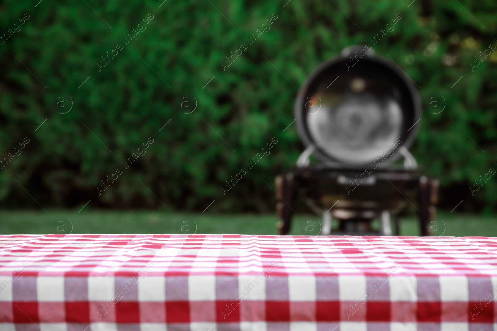 Photo of Picnic table with red checkered cloth and blurred barbecue grill outdoors