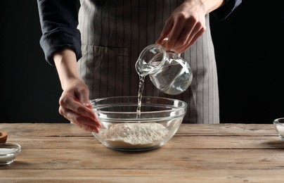 Making bread. Woman pouring water into bowl with flour at wooden table, closeup