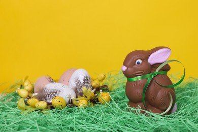 Easter celebration. Cute chocolate bunny and different eggs with feathers on grass against yellow background