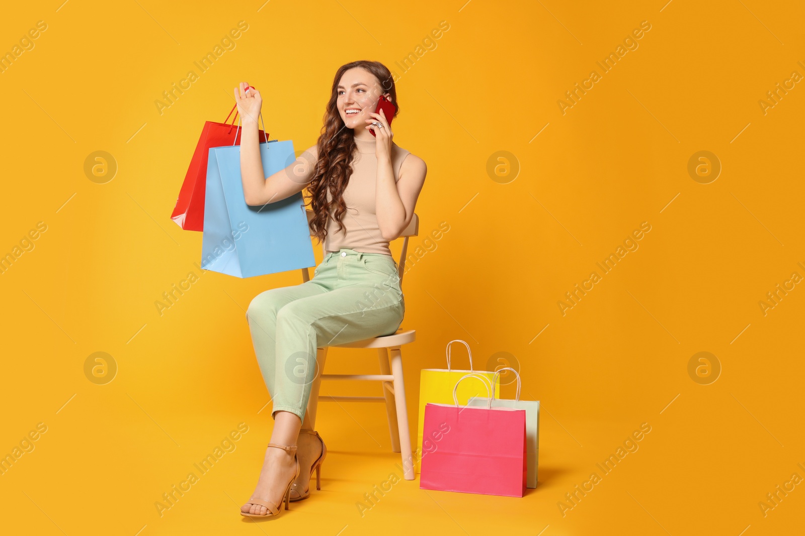 Photo of Beautiful woman holding colorful shopping bags and talking on smartphone against orange background