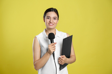 Young female journalist with microphone and clipboard on yellow background