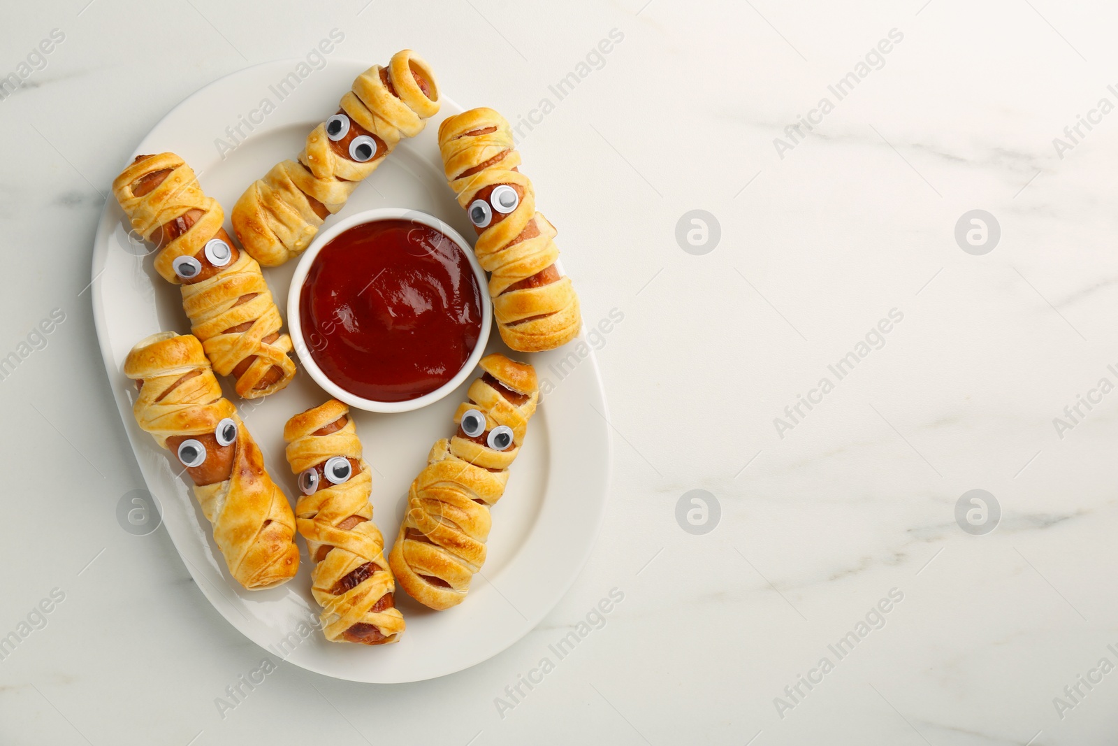 Photo of Plate with tasty sausage mummies for Halloween party and ketchup on white marble table, top view. Space for text