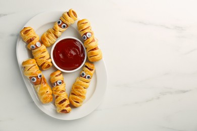 Photo of Plate with tasty sausage mummies for Halloween party and ketchup on white marble table, top view. Space for text