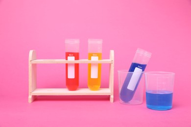 Photo of Beakers and test tubes with colorful liquids in wooden stand on bright pink background. Kids chemical experiment set