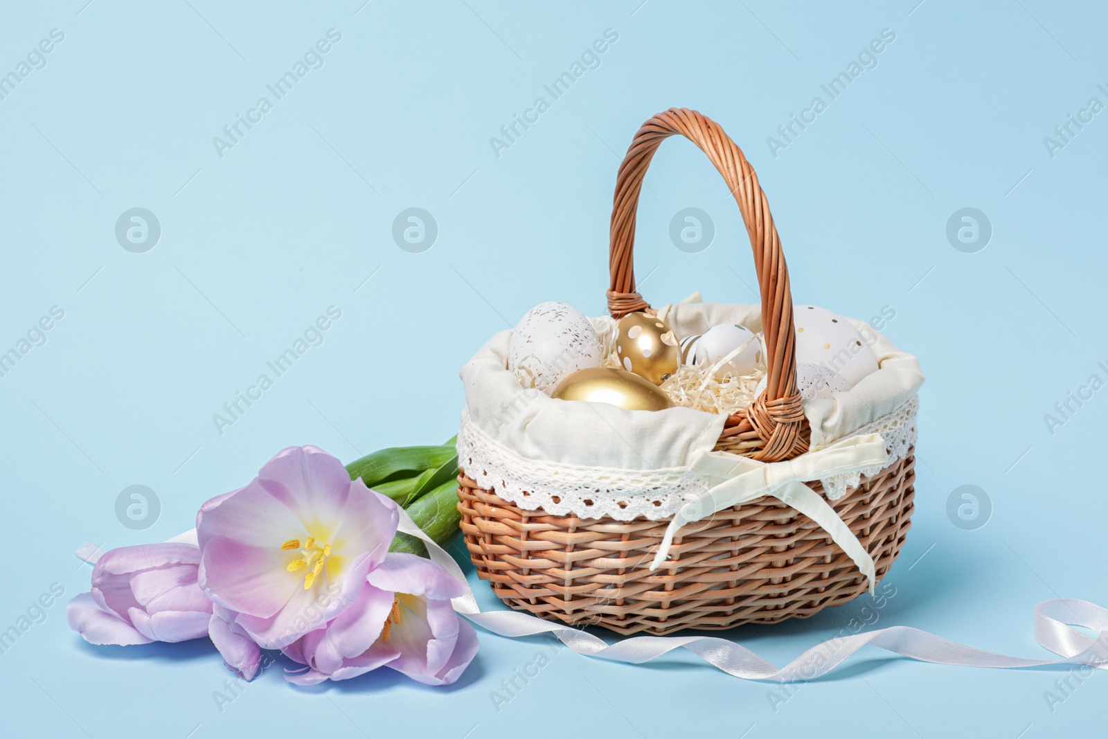 Photo of Easter basket with painted eggs, ribbon and flowers on light blue background