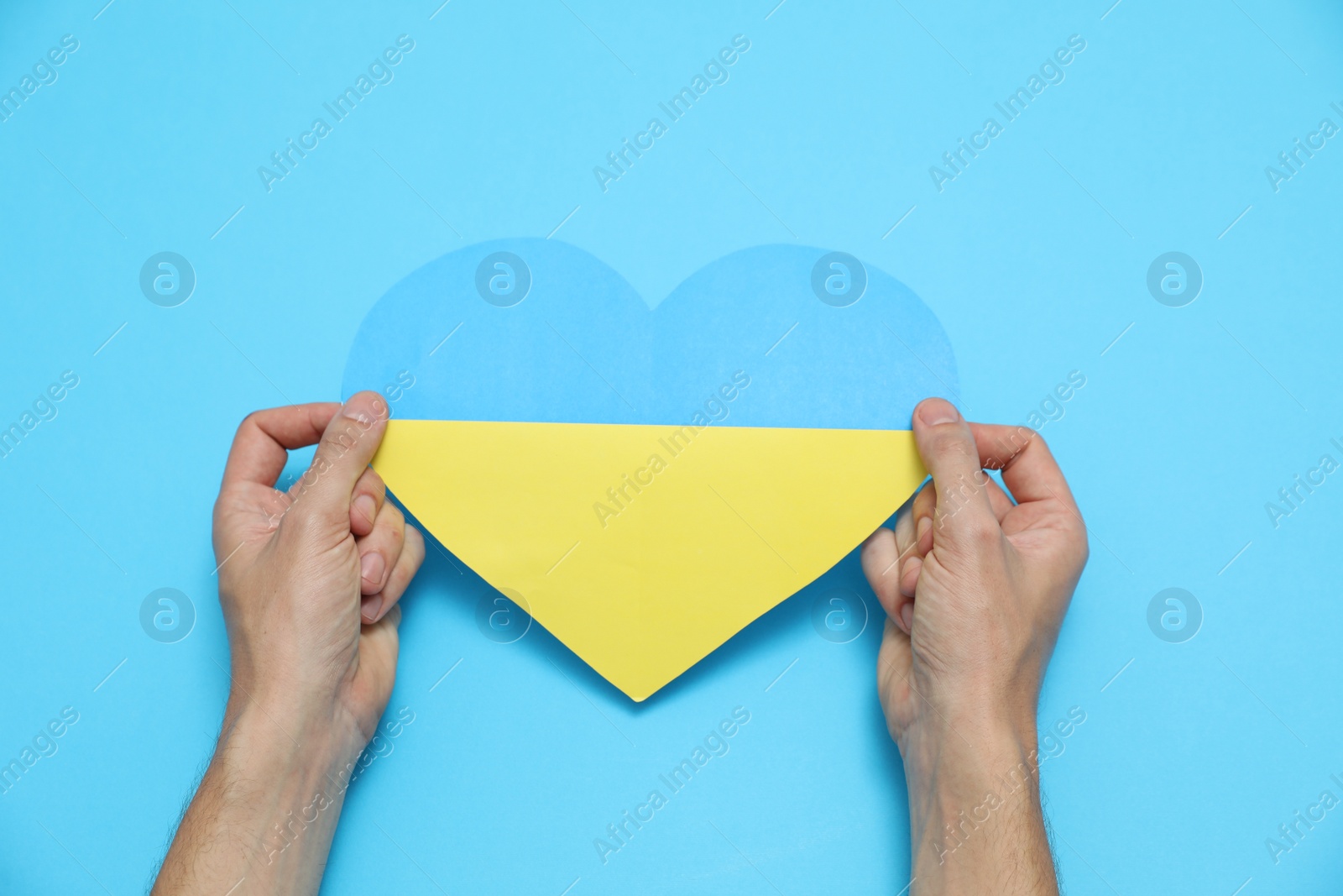 Photo of Man holding paper heart in colors of Ukrainian flag on light blue background, top view