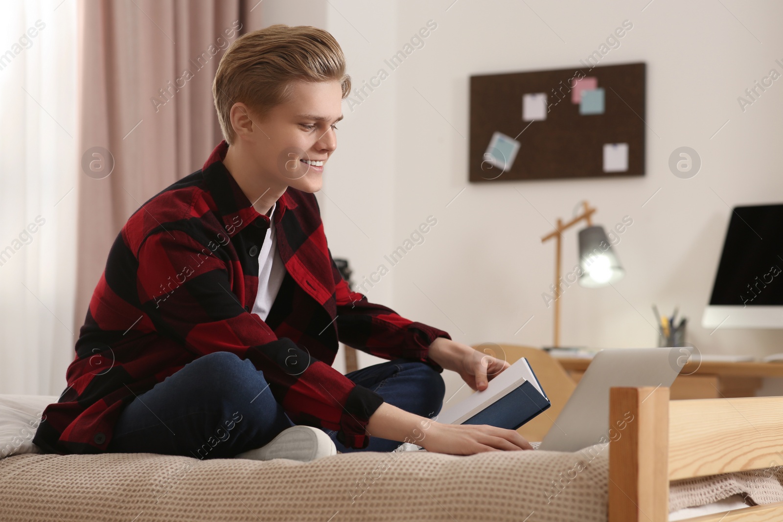 Photo of Online learning. Smiling teenage boy reading book near laptop on bed at home