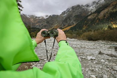 Photo of Woman holding binoculars near river in beautiful mountains, closeup. Space for text