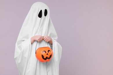 Photo of Child in white ghost costume holding pumpkin bucket on light grey background, space for text. Halloween celebration