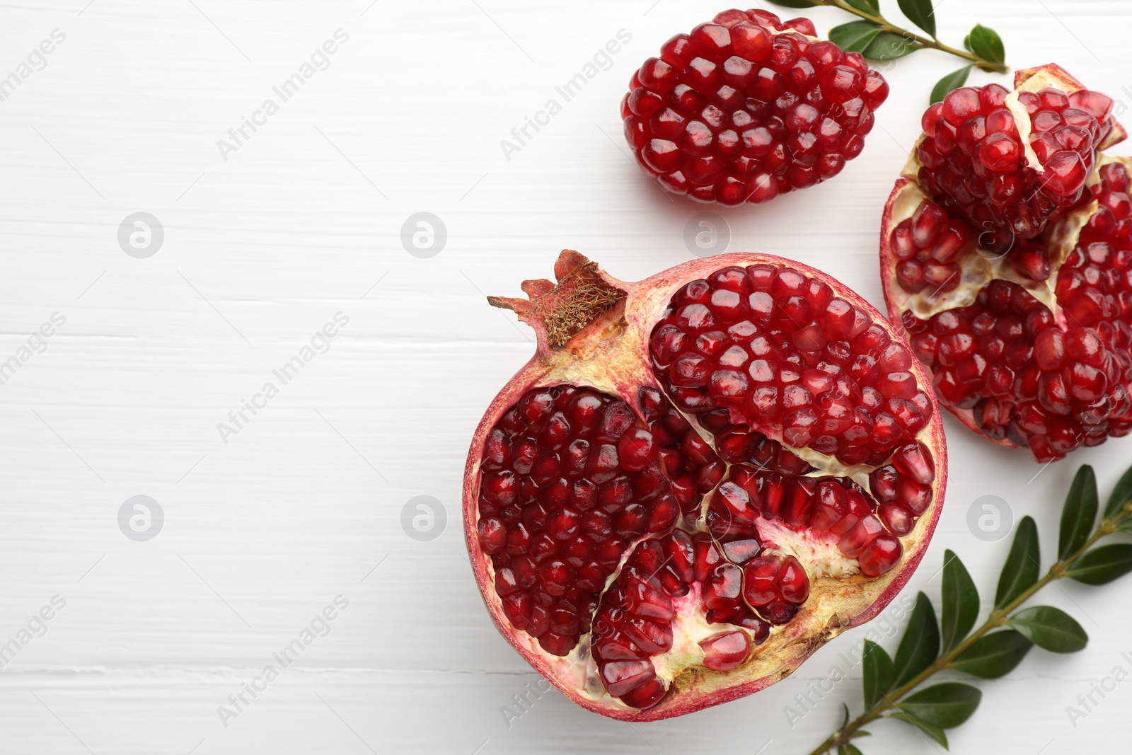 Photo of Pieces of fresh pomegranate and branches on white wooden table, flat lay. Space for text