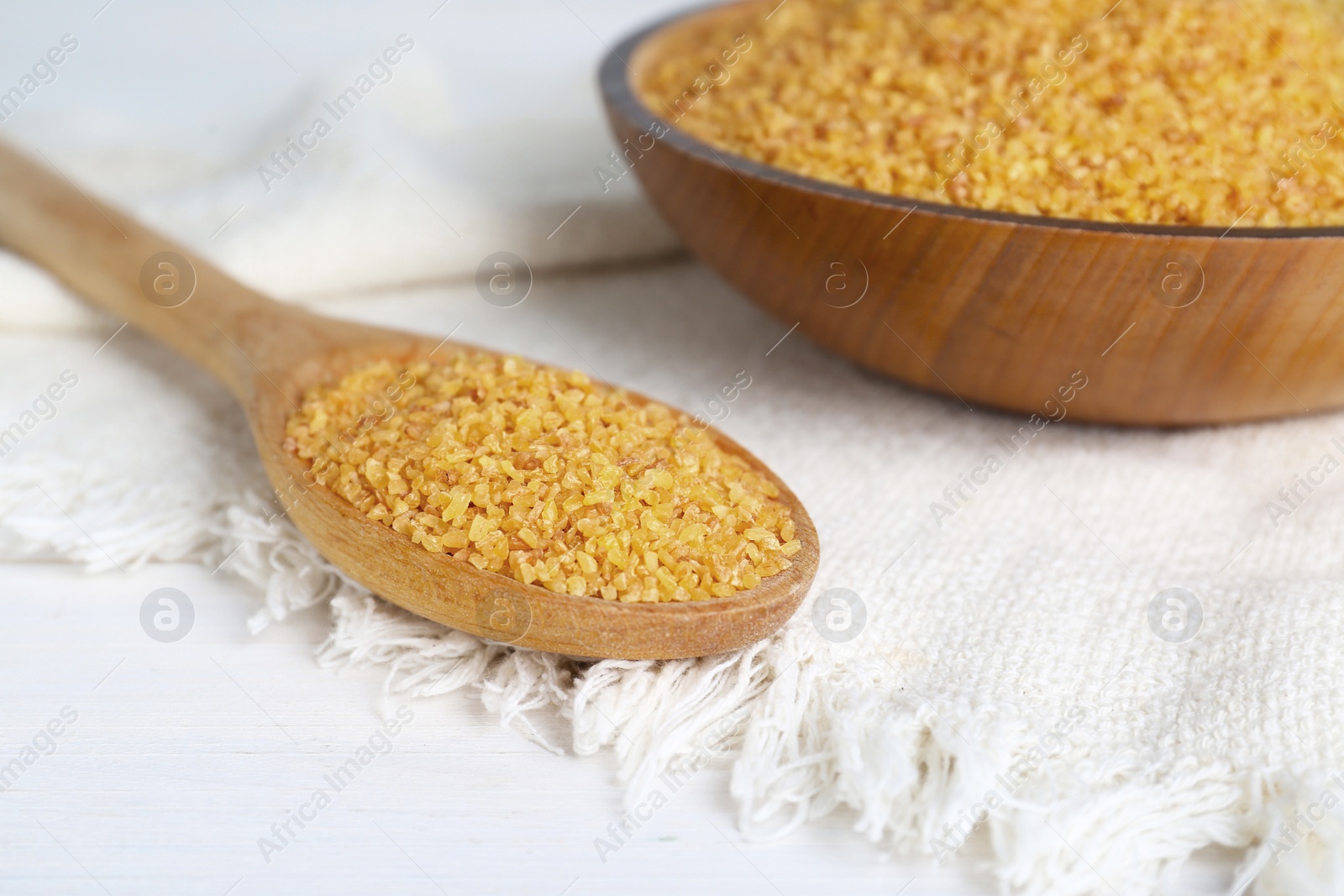 Photo of Napkin, spoon and bowl with uncooked bulgur on white wooden table, closeup