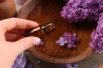 Woman pouring lilac essential oil into bowl at wooden table, closeup
