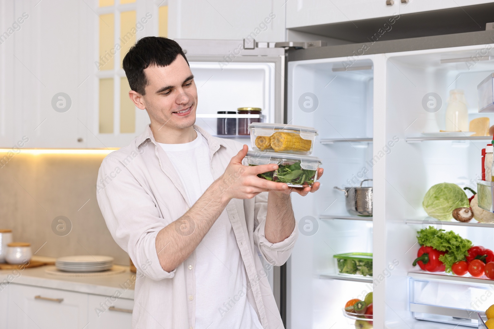 Photo of Happy man holding containers with vegetables near refrigerator in kitchen