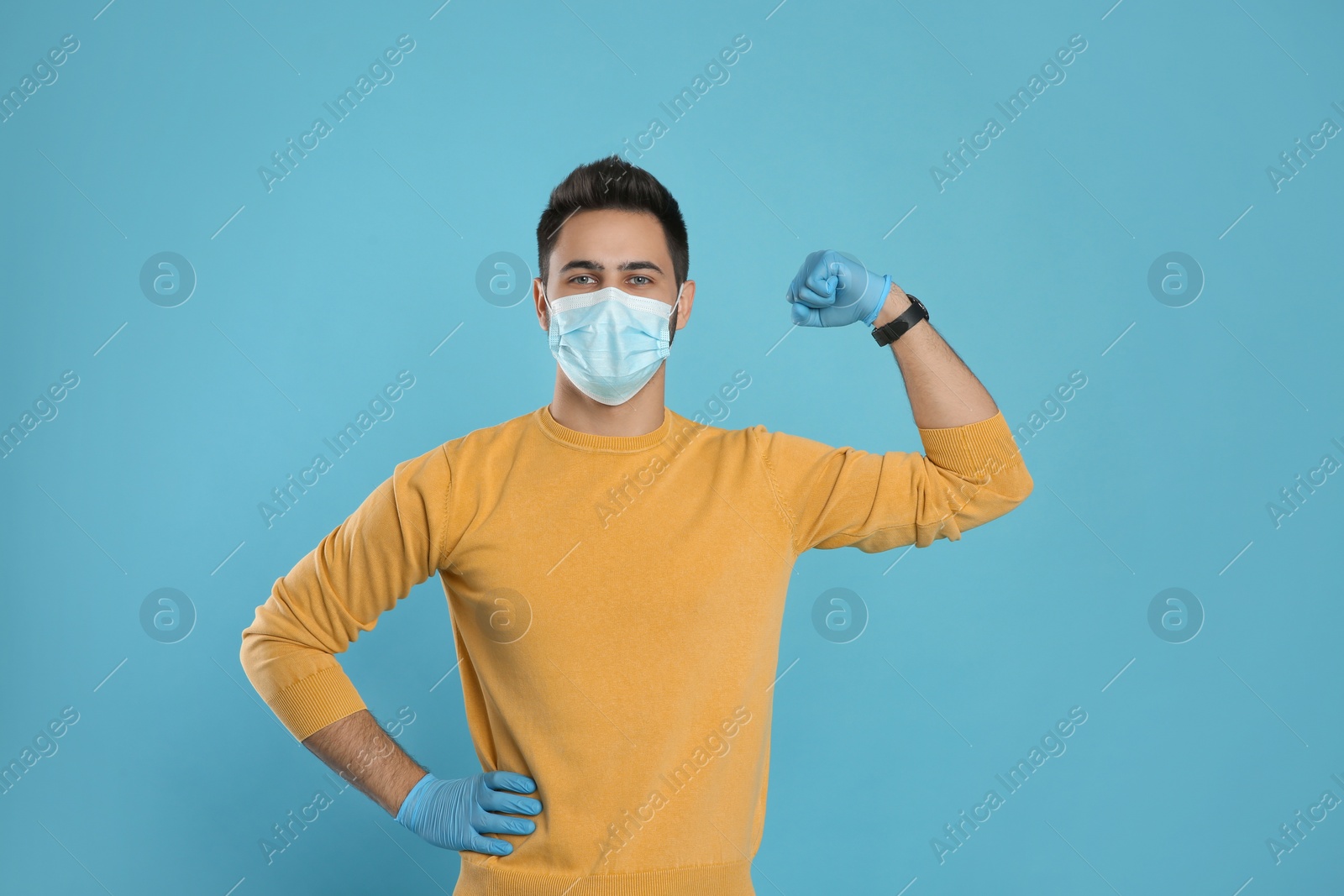 Photo of Man with protective mask and gloves showing muscles on light blue background. Strong immunity concept