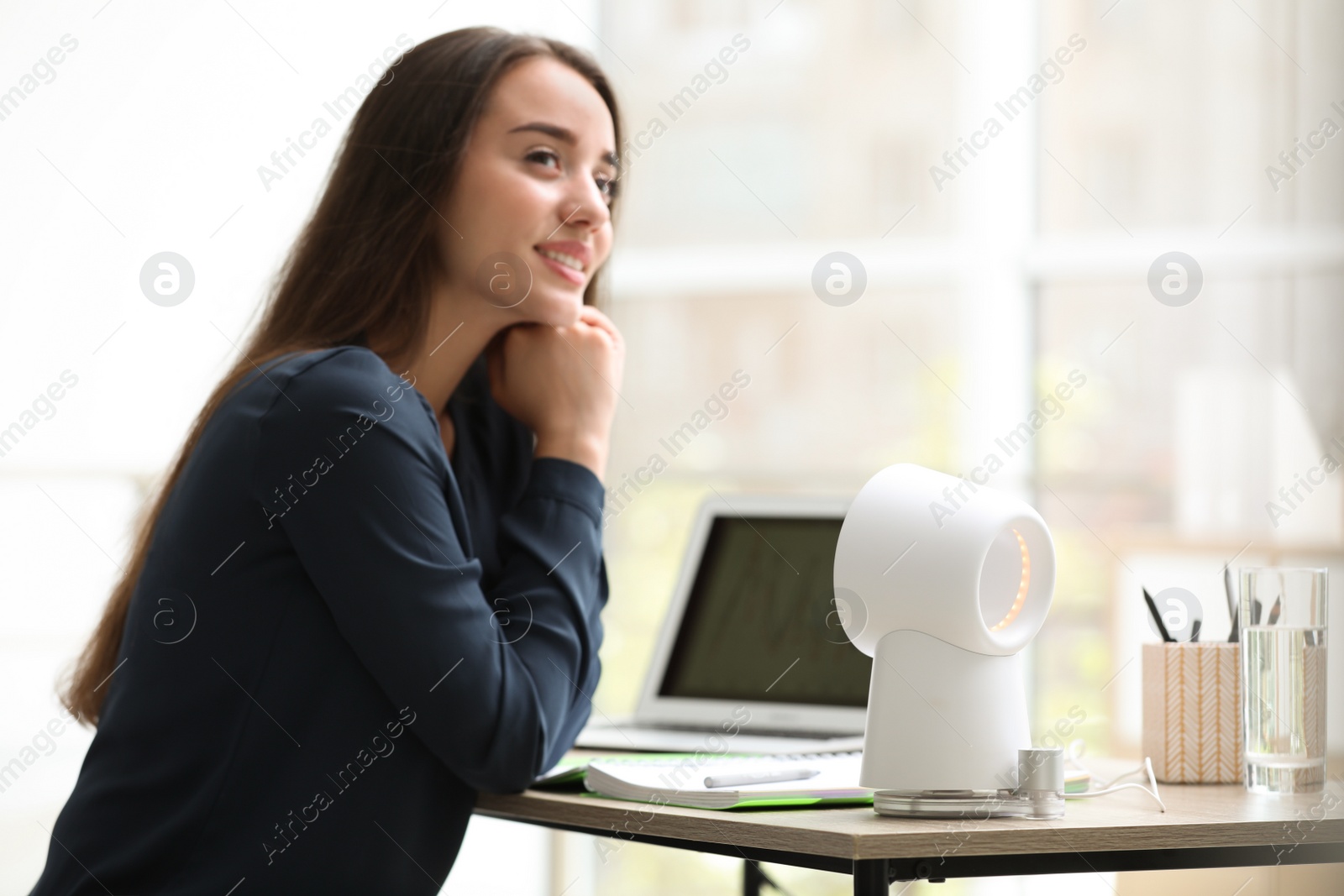 Photo of Young woman enjoying air flow from portable fan at workplace. Summer heat