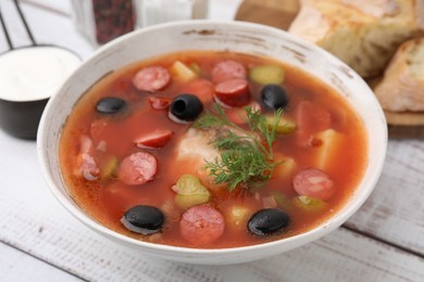 Photo of Meat solyanka soup with thin dry smoked sausages in bowl on white wooden table, closeup