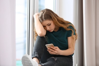 Upset woman with smartphone sitting on window sill indoors. Loneliness concept