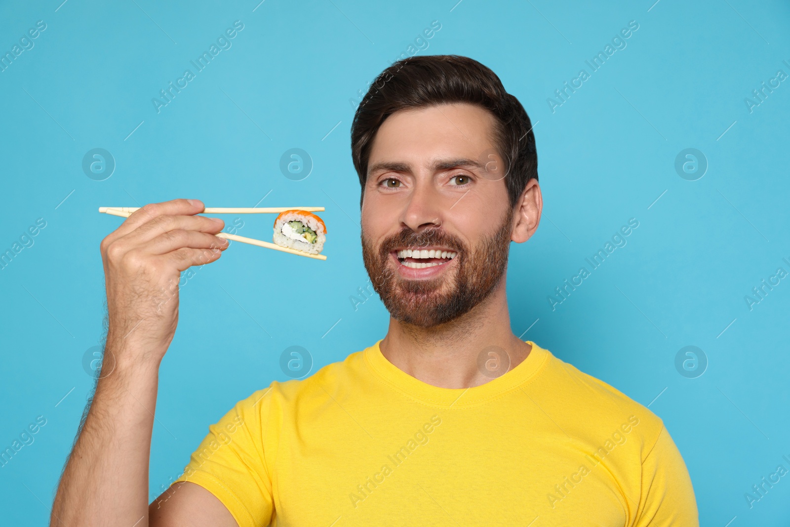 Photo of Happy man holding sushi roll with chopsticks on light blue background