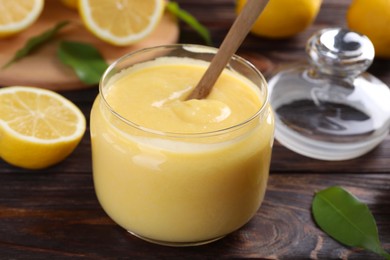 Delicious lemon curd in glass jar, fresh citrus fruits and spoon on wooden table, closeup