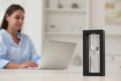 Hourglass with flowing sand on white table. Woman using laptop indoors, selective focus