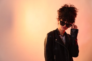 Photo of Beautiful young woman in black leather jacket and sunglasses posing on color background in neon lights. Space for text