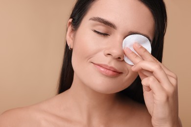 Photo of Young woman cleaning her face with cotton pad on beige background, closeup