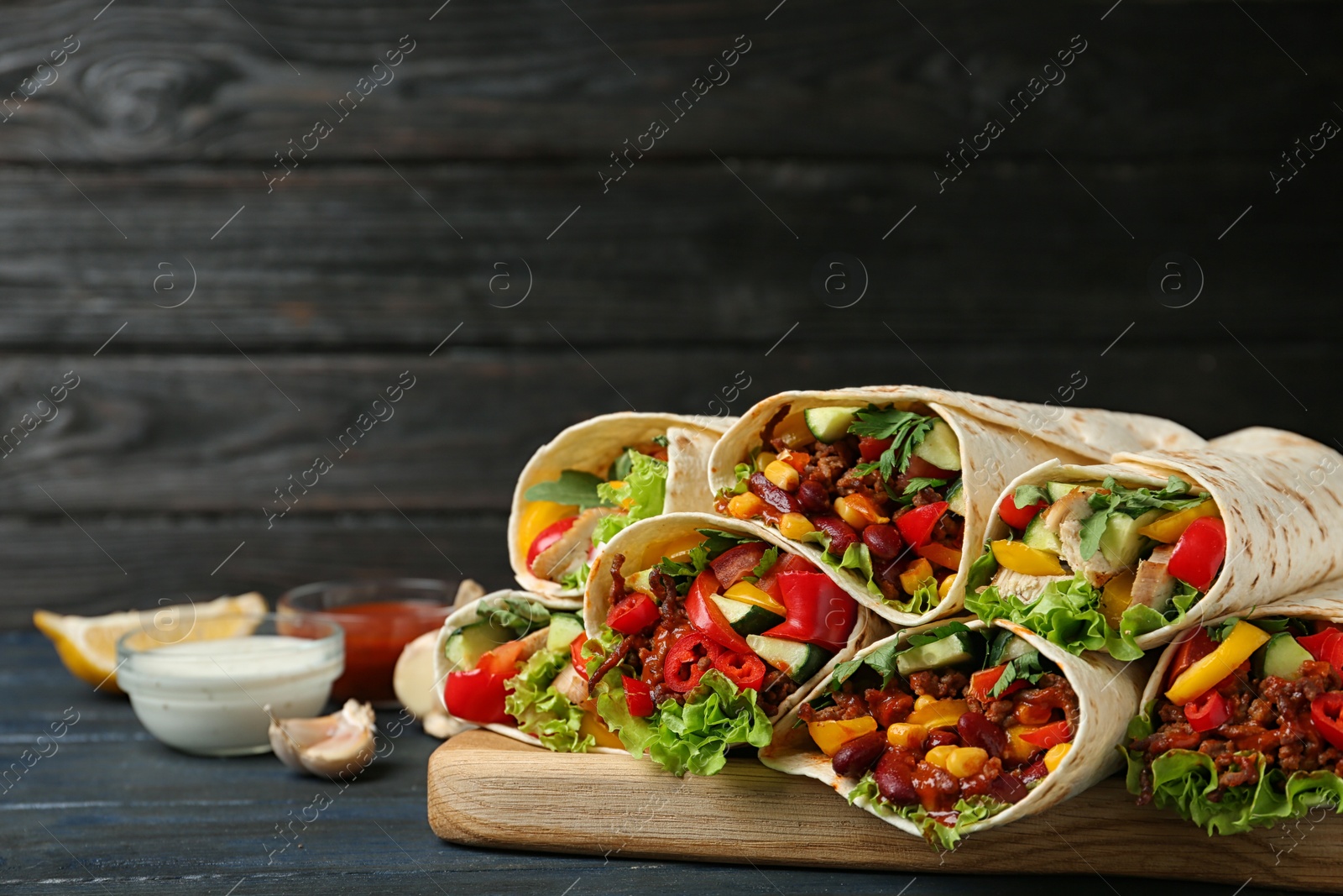 Photo of Board with delicious meat tortilla wraps on wooden table against black background. Space for text