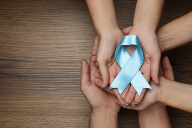 People holding light blue awareness ribbon at wooden table, top view. Space for text